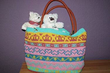 Gehäkelter bunter Shopper mit Streifenmuster und braunen Ledergriffen, zwei weiße Stofftiere darin, lila Hintergrund.