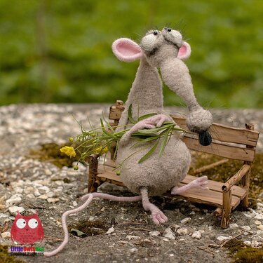 Gray crocheted amigurumi mouse with long snout and pink ears sitting on a small wooden bench holding yellow wildflowers
