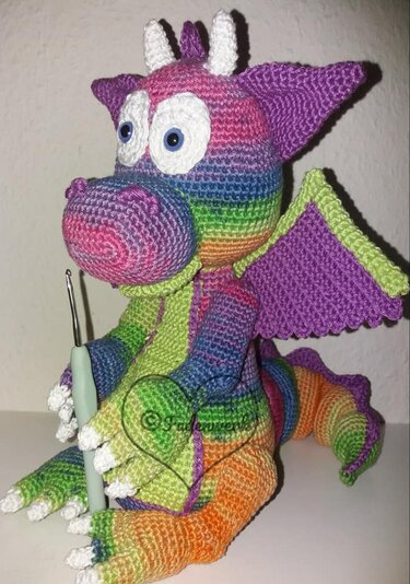 Gehäkelter Regenbogen-Drache mit großen weißen Augen, lila Flügeln und sichtbarem Häkelhaken