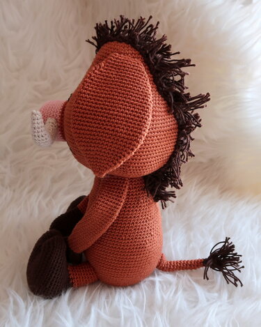 Sitzendes gehäkeltes Warzenschwein-Amigurumi in braun-orange mit dunkler Mähne und rosafarbenem Rüssel, Seitenansicht.