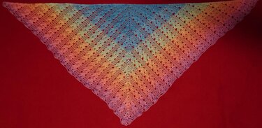Gehäkeltes dreieckiges Tuch mit Regenbogen-Farbverlauf, feinem Lochmuster und gewellter Kante auf rotem Hintergrund