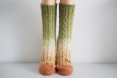 Long cable-knit socks with an ombre gradient from green to cream to rust brown and a cable pattern.