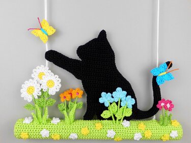 Crocheted black cat reaching for a yellow butterfly, surrounded by colorful crocheted flowers, hanging from a string.