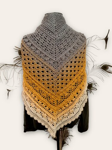 Triangle crocheted shawl on a mannequin with an ombre gradient from gray to golden yellow to cream, showing openwork stitch panels and a scalloped textured edge; feather accents around the display.