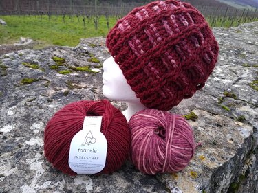 Gehäkelte burgundrote Mütze mit Rautenmuster auf weißem Schaumkopf, zwei Wollknäuel auf steinerner Mauer