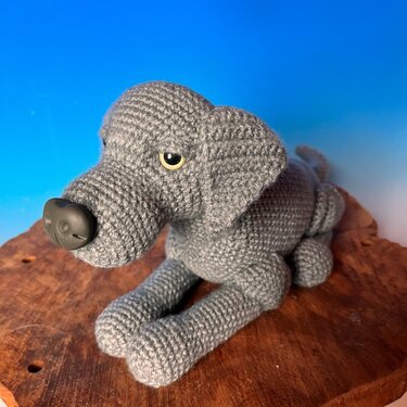 Gehäkelter grauer Amigurumi‑Hund mit gelben Sicherheitsaugen und dunkler Kunststoffnase auf Holz