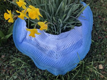 Gehäkeltes Dreieckstuch in Weiß und Blau mit Ajour- und Rippenmustern, umgeben von grünen Blättern und gelben Narzissen.