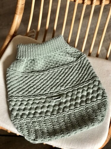 Mintgrüner gestrickter Babysack mit verschiedenen Mustern auf einem Kissen in einem Rattanstuhl