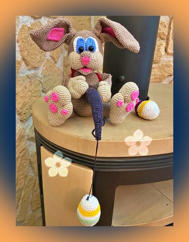 Brauner gehäkelter Amigurumi-Osterhase mit blauen Augen und rosa Akzenten auf einem Holzsockel, umgeben von gehäkelten, gestreiften Ostereiern.