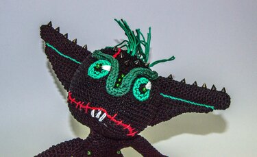 Schwarzes gehäkeltes Amigurumi-Monster mit grünen Augen, roten Sticknähten und Spikes an den Ohren.