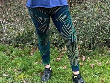 Beine in gestrickten blau-grünen Leggings mit geometrischem Linienmuster und schwarzen Schuhen, stehend auf Gras