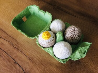 Grüner Eierkarton mit drei runden und einem Spiegelei-ähnlichen gehäkelten Ei auf einer hellen Holzfläche.