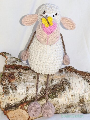 Weißes gehäkeltes Schaf-Amigurumi mit rosa Ohren und Schnauze und gelbem Schmetterling auf der Nase, sitzend auf einem Birkenholzstamm.