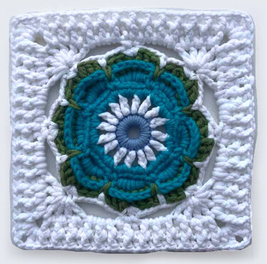 Nahaufnahme eines quadratischen Häkelmotivs mit Blumenmuster in Blau, Türkis, Weiß und Grün, umgeben von weißem Rand.
