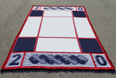 Rechteckige Strickdecke in Weiß, Dunkelblau und Rot mit Gitterfeldern, Zopfmuster an den kurzen Seiten, roten Fransen und eingestrickten Zahlen "20".