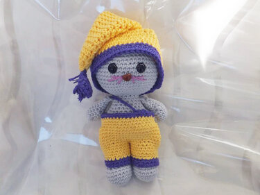 Gehäkelte graue Amigurumi-Maus mit gelber Zipfelmütze und gelber Latzhose, lila Details, auf hellem Hintergrund.