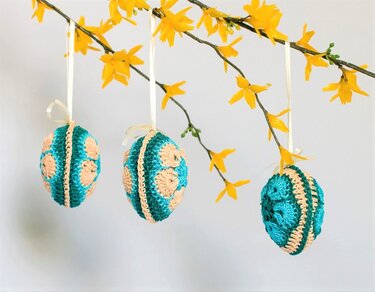 Drei gehäkelte Ostereier im African-Flower‑Motiv in Türkis und Beige an einem blühenden Forsythienzweig.