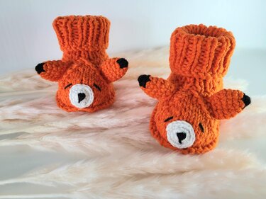 Orange gestrickte Babyschuhe mit Fuchs-Gesicht auf hellem, flauschigem Untergrund