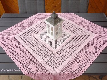 Rosafarbener gehäkelter Tischläufer mit Herz- und Filetmuster, diagonal auf einem dunklen Tisch mit weißer Laterne.