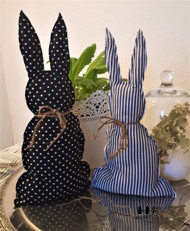 Zwei genähte Stoffhasen, einer schwarz mit weißen Punkten, einer schwarz-weiß gestreift, jeweils mit Juteschleife auf Tablett neben Pflanze