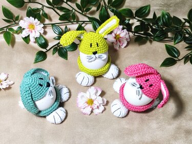 Drei gehäkelte Hasen-Eierwärmer in Grün, Türkis und Pink auf beigem Stoff, dekoriert mit Blüten und grünen Blättern.