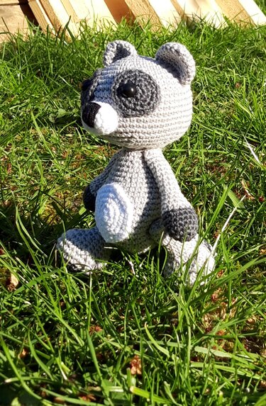 Gehäkeltes graues Waschbär-Amigurumi mit weißem Bauch sitzt im Gras