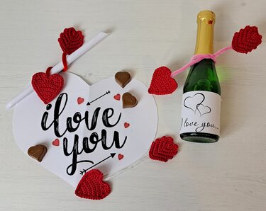 Gehäkelte rote Herzen und Rosen neben einer Herzkarte mit 'I love you', Schokoladenherzen und einer dekorierten Flasche auf hellem Untergrund.