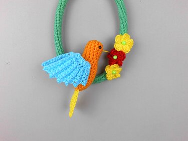 Gehäkelter Kolibri mit orangefarbenem Körper und hellblauen Flügeln an einem grünen Häkelkranz mit gelben und roten Blumen und grünen Perlen.