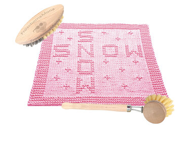 Zartrosa gestrickter Topflappen mit 'SNOW'-Muster und dekorativer Randbordüre, neben zwei Holzbürsten auf weißem Hintergrund