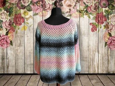 Gehäkelter Damenpullover in Pastellrosa, Hellblau und Grau mit Farbverlauf auf schwarzer Schneiderpuppe vor hölzernem, floralem Hintergrund