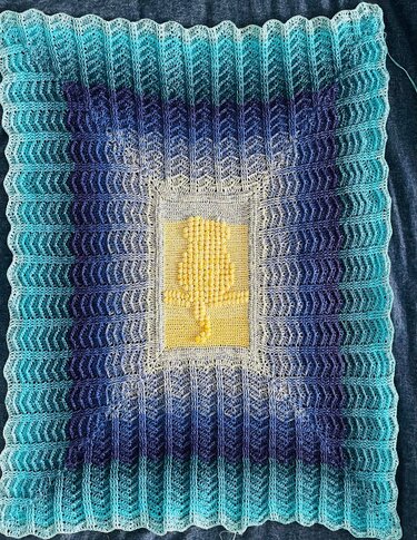 Gehäkelte Decke mit türkis-blauem Farbverlauf und gelbem, reliefartigen Bobble-Mittelteil