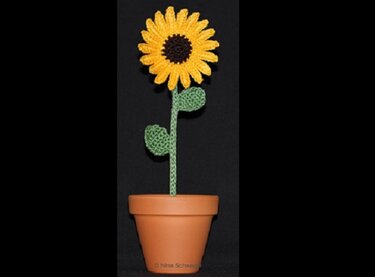 Gehäkelte gelbe Sonnenblume mit grünem Stiel in Terrakottatopf vor schwarzem Hintergrund.
