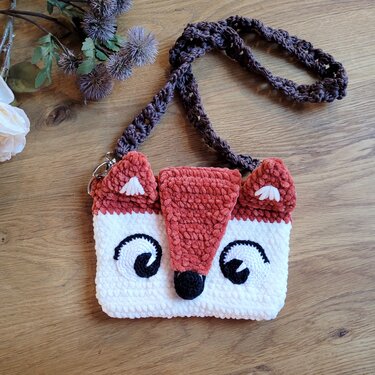 Flauschige weiß-rostbraune Häkeltasche mit Fuchsgesicht und dunkelbraunem Trageriemen auf einem Holztisch.