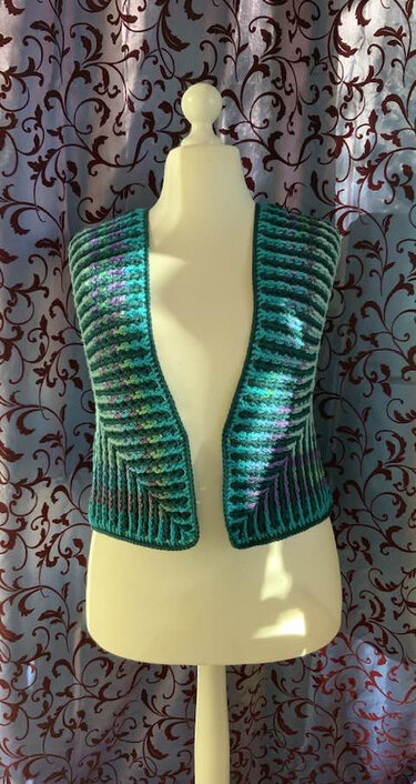 Türkis-grüner gehäkelter Bolero mit Rippenmuster auf Schneiderpuppe vor gemustertem Vorhang