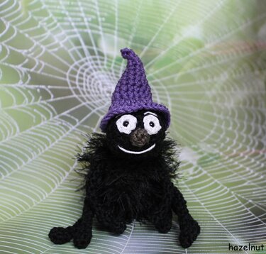Schwarze Amigurumi-Spinne mit lila Hexenhut vor Spinnennetz