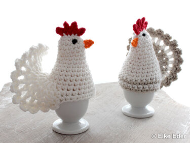 Zwei gehäkelte Hühner-Eierwärmer in Weiß und Beige auf weißen Eierbechern