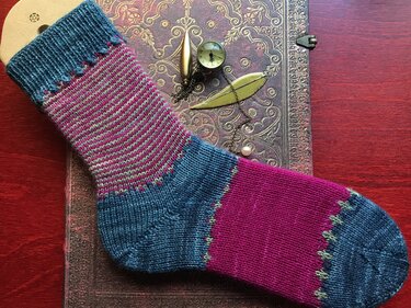Gestrickte Socke in Petrol und Magenta mit kontrastfarbener Ferse und Spitze auf verziertem Notizbuch