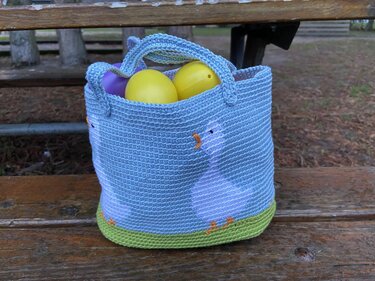 Hellblauer gehäkelter Korb mit Gänsemotiven, grünem Boden und gelben Plastikeiern