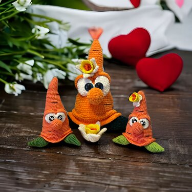 Drei orange gehäkelte Karotten-Amigurumi mit Blüten, grünen Blattfüßen und großen Augen auf einem Holztisch; im Hintergrund rote Herzdekoration