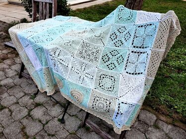 Gehäkelte Decke aus Granny-Squares in Pastelltönen auf einer Holzbank im Garten