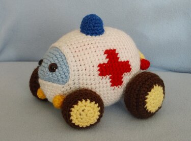 Gehäkelter Amigurumi-Krankenwagen, weiß mit rotem Kreuz, braune Räder und blaues Blaulicht