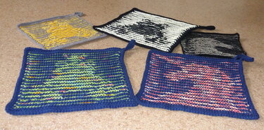 Fünf quadratische, gestrickte Topflappen in verschiedenen Farben mit Aufhängeschlaufen, liegend auf dem Boden