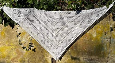 Weißes handgehäkeltes Dreieckstuch mit Herz-Lochmuster, aufgehängt vor einer Efeuwand.