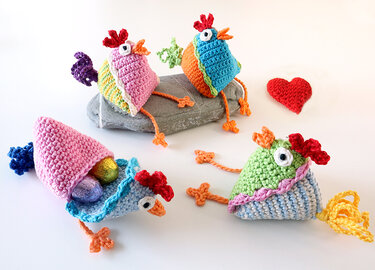 Four bright crocheted amigurumi chickens, one used as an egg cozy holding foil-wrapped eggs, with a small red crocheted heart.