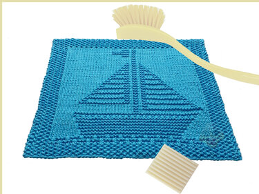 Türkis gestricktes quadratisches Spültuch mit eingestricktem Segelbootmotiv und strukturiertem Rand; daneben eine Spülbürste und ein Stück Seife.