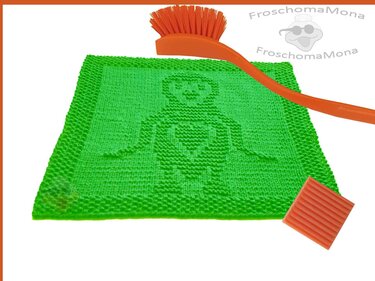 Quadratisches grünes gestricktes Spültuch mit eingestrickter Personenfigur und Herzmotiv, dazu orangefarbene Bürste und Schwamm