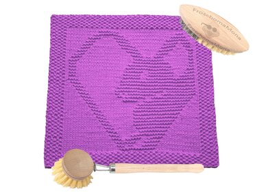 Lila gestricktes quadratisches Tuch mit eingearbeitetem Herzmotiv und zwei hölzernen Bürsten auf weißem Hintergrund.