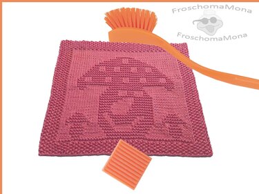 Rosa gestricktes Spültuch mit Pilzmotiv, orangefarbene Spülbürste und Seifenstück auf hellem Hintergrund.