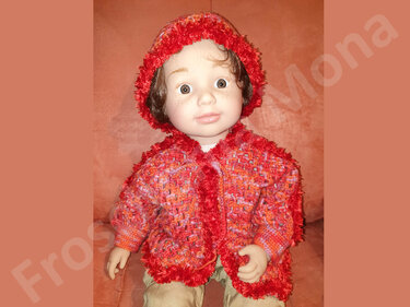 Babypuppe trägt eine gehäkelte, flauschige Jacke und Kapuze in Rot- und Orangetönen vor braunem Hintergrund.