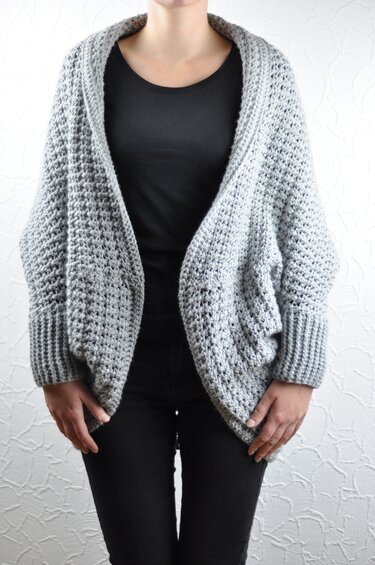 Person wearing a light grey chunky crochet shrug with ribbed cuffs over a black top
